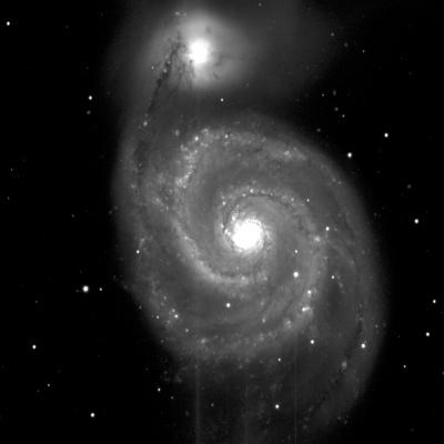 A large spiral galaxy in shades of grey. It has a bright, white centre, with dusty, grey arms swirling around it. At the centre-top is another bright, white circle with some light grey fuzziness around it. The background is black with small, white dots of stars.