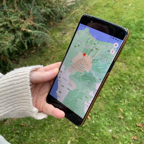 A hand holding a smartphone displaying a map with a red location pin, against a background of green grass and plants