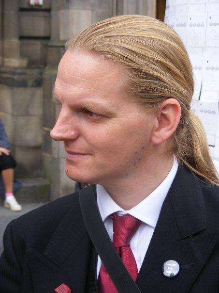 A portrait photograph of a man looking towards the image left, his face slightly angled that way. He has long, blond-brown hair tied back into a low ponytail, and he is wearing a black suit with a red tie.