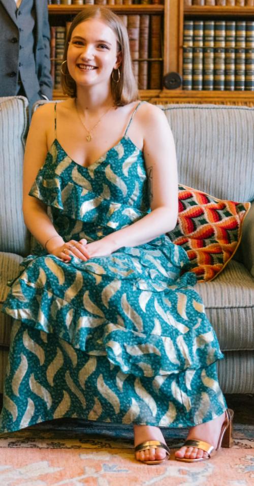 A woman with shoulder length, dark blonde hair sat down on a couch. She is wearing a green dress with gold waves of pattern. She is smiling and facing towards the camera.