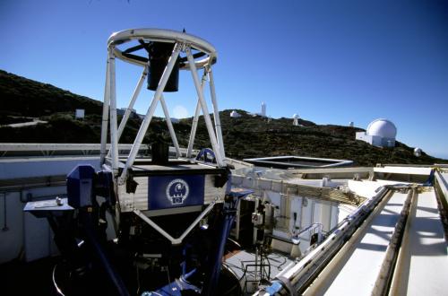 A close-up view of the Liverpool Telescope. It is pointing upwards at the sky which is bright blue in colour. The telescope is within an enclosure with its roof open and folded back. In the distance, there are a few more white telescope enclosures on a sloping hill.