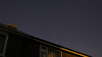 A night sky filled with stars above the roof of a house. The house is partially illuminated with a warm indoor light and a purple glow from one of the windows. The clear sky shows numerous stars twinkling above