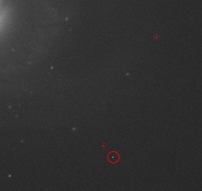 A fuzzy black/dark grey background with a few light grey spheres randomly throughout. Towards the upper left, there is a faint light grey region, as if something bright is beyond the image. In the upper right and centre-bottom, there are in total three tiny, white streaks which are circled in a red outline.